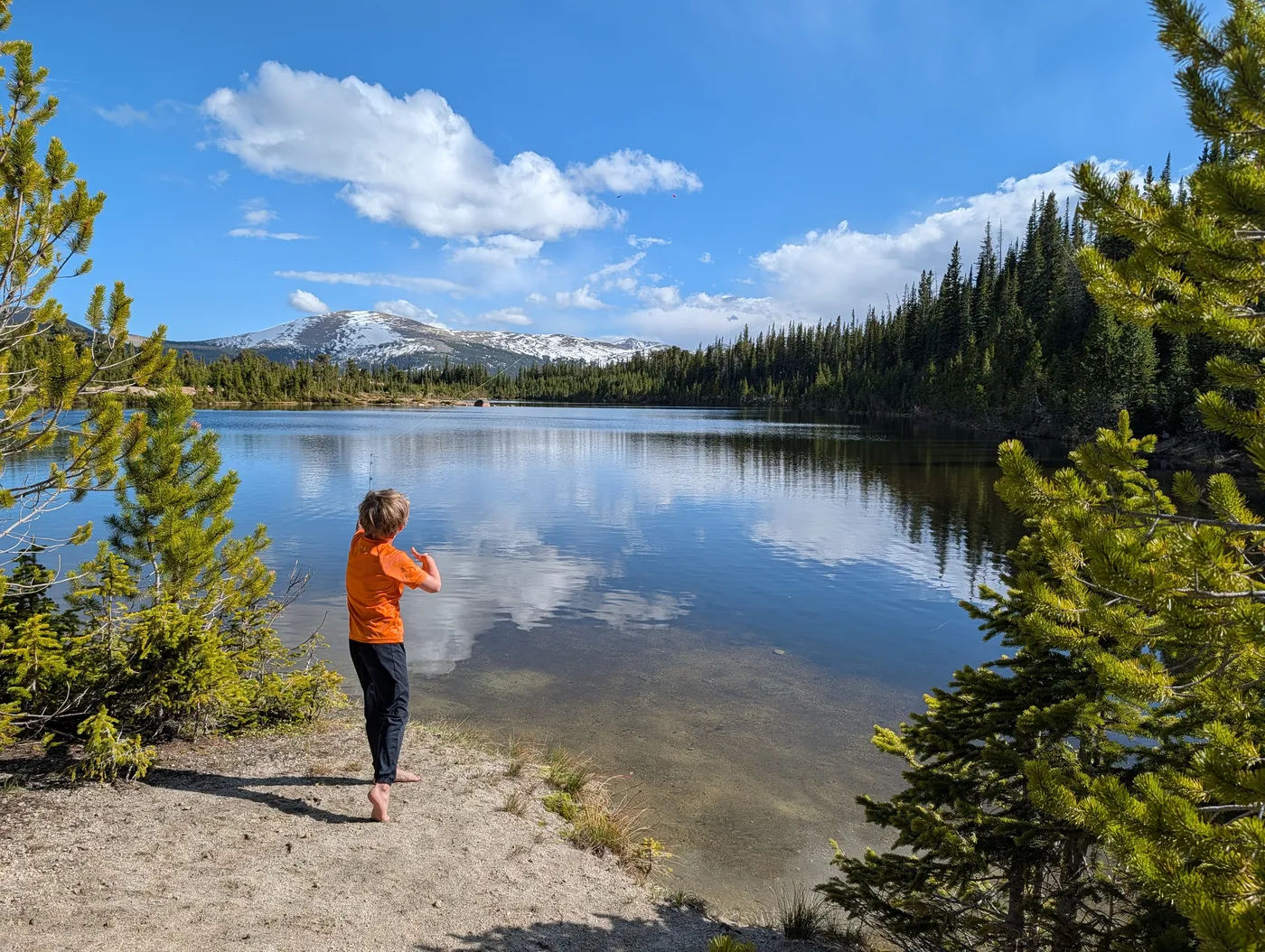 A barefoot boy in an orange shirt skipping stones at a calm mountain lake, snowy peaks and pine trees beyond