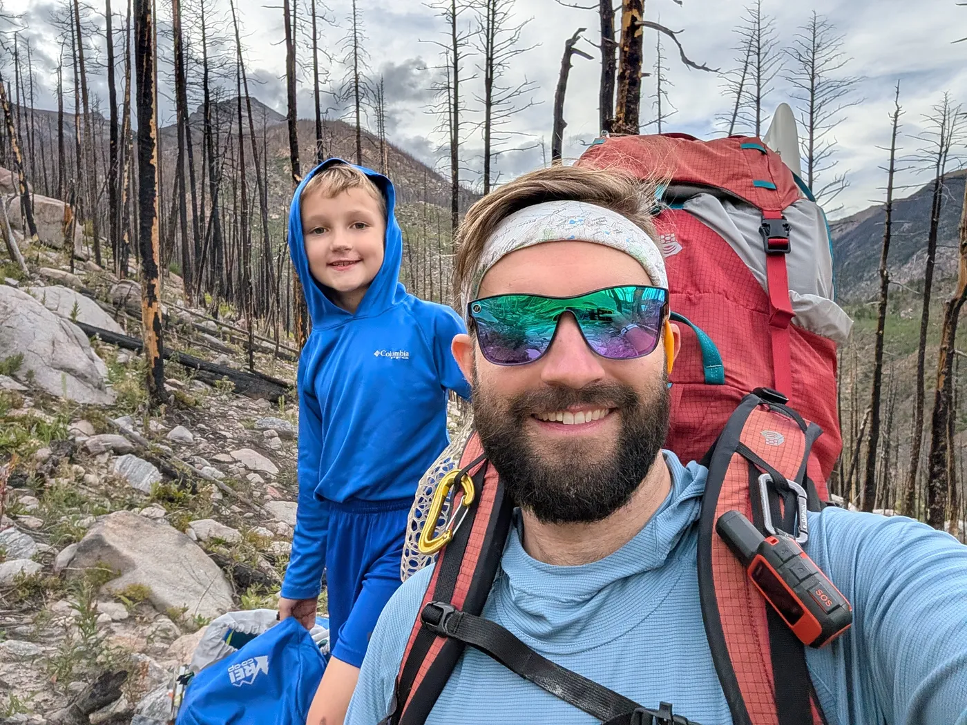 A dad and his young son smiling on a rocky mountain trail through a burned forest, the dad carrying a big red backpack